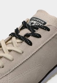 Κοντινό πλάνο μπεζ σουέντ αθλητικού παπουτσιού Converse με ανάμεικτα μαύρα και κρεμ κορδόνια και εμφανείς ραμμένες λεπτομέρειες.