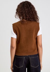 Suéter marrón de punto sin mangas con dobladillo acanalado, combinado con una camisa blanca de manga corta, llevado con jeans de mezclilla oscura.