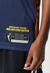 Μπλε διχτυωτή μπασκετική φανέ λα με την επιγραφή «Indiana Fever» και «Nike Explorer Edition» σε κίτρινο χρώμα. Περιλαμβάνει μια μαύρη και λευκή ετικέτα με το μέγεθος και το λογότυπο.