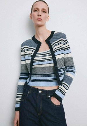 Mujer con un cárdigan de rayas acanalado en azul, blanco y negro sobre un top corto a juego, combinado con vaqueros azul oscuro, con la mano en el bolsillo.