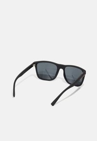 Ochelari de soare din plastic negru cu lentile gri, formă pătrată și temple elegante. Au un accent mic din metal lângă balama.