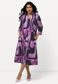 Robe violette arborant un motif géométrique dans des teintes plus sombres, avec des manches trois-quarts, un décolleté en V et des poches latérales. Associée avec des talons roses.