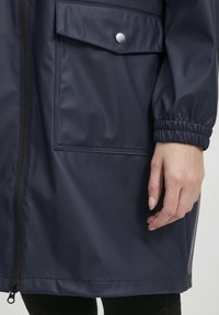 Veste imperméable marine avec fermeture à zip, poignets élastiques et poche avant avec bouton-pression. Texture lisse et brillante.