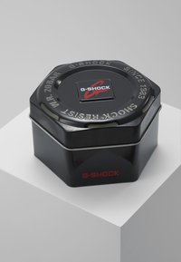 Conteneur en métal hexagonal noir avec une finition lisse, emblématique d'un logo G-Shock rouge et d'un texte indiquant la résistance aux chocs et à l'eau.
