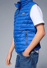 Blå vadderad väst med hög krage, utrustad med dragkedja och sidofickor. Bärs över en vit t-shirt, kombinerad med denimjeans.