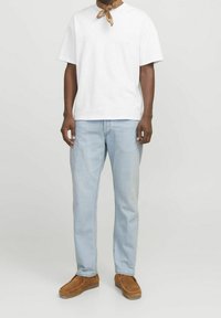 Camiseta de algodón blanca con cuello redondo, combinada con jeans de mezclilla azul claro. El modelo lleva zapatos tipo slip-on de ante marrón y una bufanda marrón.