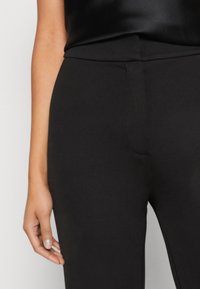 Leggings noirs taille haute en tissu lisse et extensible. Comprend une ceinture plate et une silhouette ajustée, avec un détail de couture visible sur la poche.