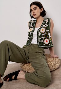 Gilet di maglia con ricami floreali verdi, chiusura con bottoni, indossato su una canotta bianca; abbinato a pantaloni larghi verde oliva e sandali neri.