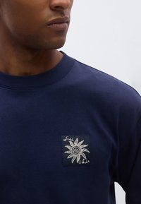 Φούτερ σε ναυτικό μπλε με στρογγυλή λαιμόκοψη και ένα μικρό διακοσμητικό πατς που απεικονίζει έναν λευκό ήλιο και το όνομα της μάρκας. Απαλό βαμβακερό υλικό.