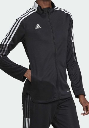 Osoba ubrana w czarną kurtkę i spodnie sportowe Adidas z białymi paskami i logo, stoi z jedną ręką w kieszeni, a drugą luźno opartą.