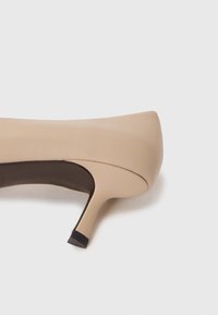 Pump beige con tacco alto, realizzata in pelle liscia, con punta affusolata e tacco sottile. La suola presenta un accento in gomma nera a contrasto.