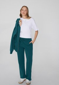 Teal Sweatpants mit elastischem Bund und seitlichen Taschen, kombiniert mit einem weißen T-Shirt. Das Model hält eine passende tealfarbene Jacke über der Schulter.