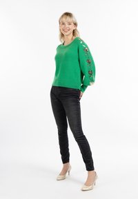 Maglione verde lavorato a maglia con paillettes a forma di cuore sulle maniche, abbinato a jeans neri aderenti e scarpe con tacco dorate.
