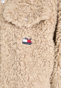 Beige fleecejakke med en teksturert, krøllete overflate og et lite blått, hvitt og rødt logo-patch som står "TOMMY JEANS" på forsiden.