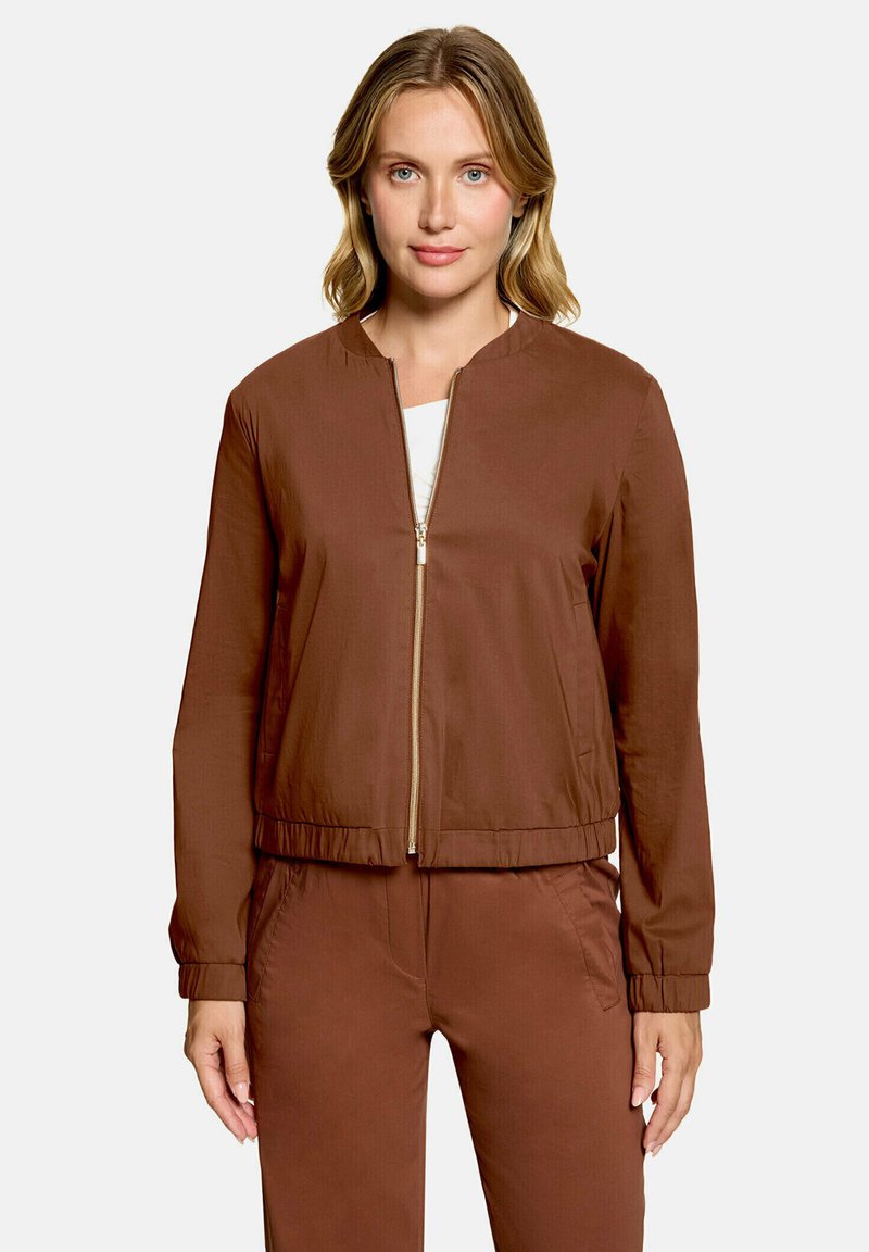 Femme aux cheveux blonds portant une veste marron zippée et un pantalon marron assorti, debout devant un fond gris clair uni.