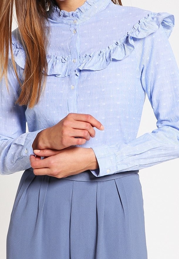 Femme portant un chemisier bleu clair boutonné avec des volants et des pois subtils, associé à un pantalon bleu plissé taille haute.
