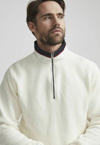 Witte textuurpullover met een ritskraag. Heeft een marineblauwe kraag met rode accenten. Het ontwerp is modern en casual.
