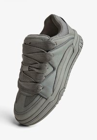 Stevige grijze sneaker met dikke geweven veters, gewatteerde kraag en gestructureerde zool, schuin geplaatst op een witte achtergrond.