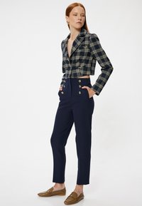 Kurz geschnittenes Blazer in Marineblau und Beige mit einem Spitzrevers, kombiniert mit hoch geschnittenen Marineblau-Hosen mit goldenen Knopfakzenten. Braune Slipper.