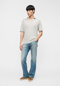 Νεαρός άνδρας στέκεται με το ένα χέρι στην τσέπη, φορώντας ανοιχτό γκρι πλεκτή κοντομάνικη μπλούζα, μπλε τζιν και καφέ δερμάτινα αθλητικά παπούτσια, μπροστά σε απλό φόντο.