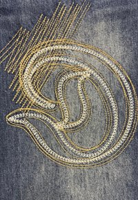 Diseño bordado en denim oscuro, que presenta una forma fluida en hilos blancos y dorados con patrones de puntada y texturas variadas.