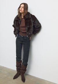 Ruskea tekoturkistakki, joka on lyhyt, puetaan istuvan ruskean neuleen päälle. Yhdessä mustien kapeiden farkkujen ja ruskeiden löysien nilkkureiden kanssa.