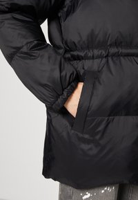 Doudoune noire avec taille cintrée, poignets élastiques, une poche latérale et une texture de tissu lisse et brillante.