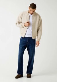 Beżowa dzianinowa kurtka bomberka z prążkowanymi mankietami, noszona na białej koszuli, w połączeniu z ciemnoniebieskimi jeansami i brązowymi butami.