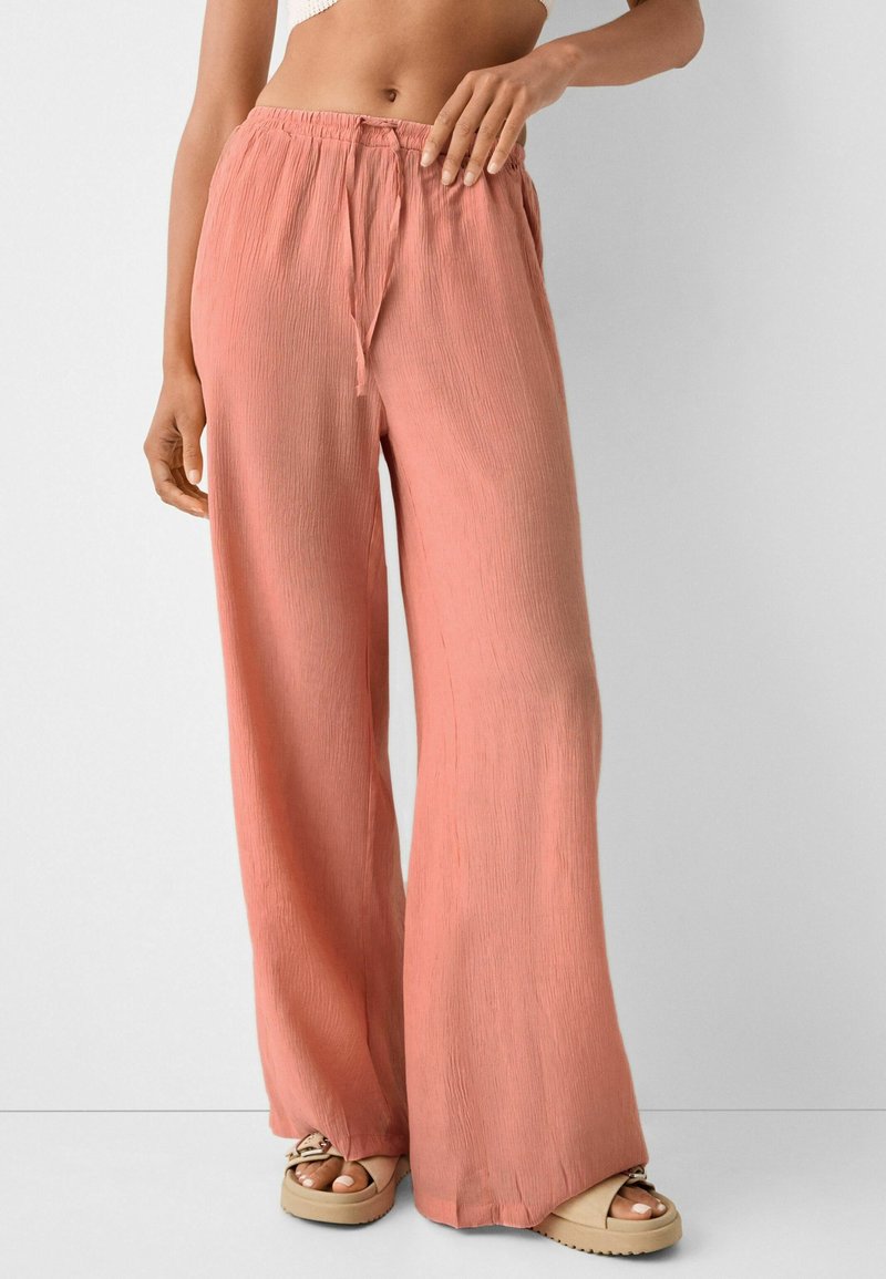 Femme portant un pantalon large rose froissé et ample avec une taille élastique et un cordon de serrage, assorti à des sandales à plateforme beiges.