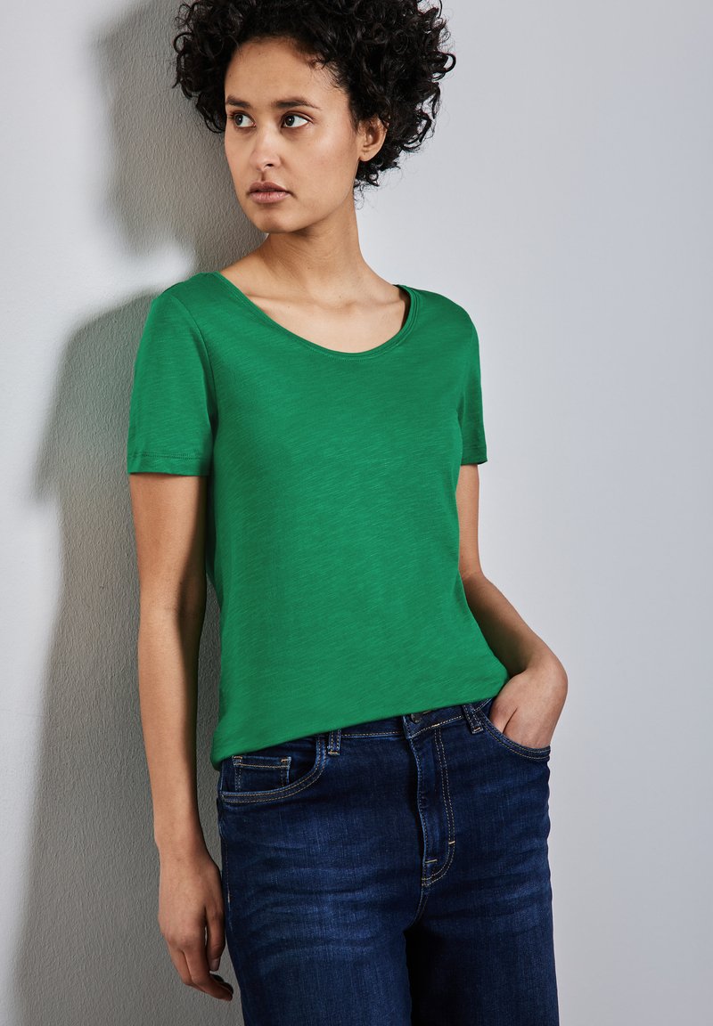 Zelené tričko s krátkým rukávem a výstřihem do véčka, vyrobené z lehkého materiálu; v kombinaci s tmavě modrými džíny, které mají volný střih.