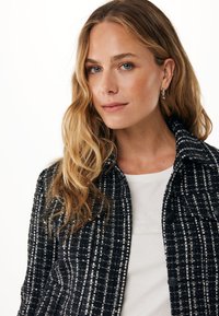 Giacca in tweed bianco e nero intrecciato con collo a punte e chiusure a bottone, indossata sopra una camicia bianca con sottili decorazioni.
