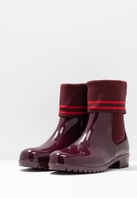 Botas de lluvia de tobillo de un rojo oscuro brillante con puños de punto ribeteados gruesos doblados hacia abajo, que presentan dos franjas rojas horizontales en cada puño.