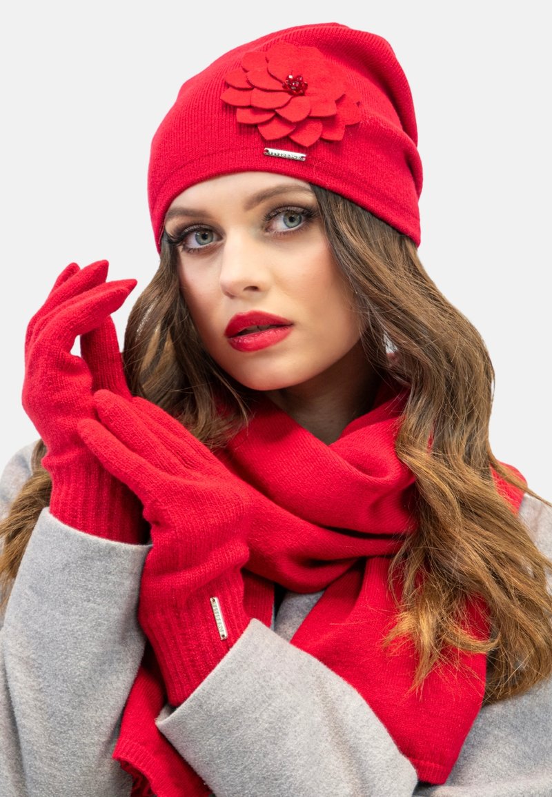 Bonnet tricoté rouge avec un accent en feutre en forme de fleur, gants rouges assortis et écharpe rouge. Texture douce, design ajusté et détails matériels simples.