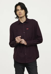 Bordeaux languit-shirt met een button-down kraag; heeft een borstzak en een subtiel merkembleem; stof lijkt textuur te hebben.