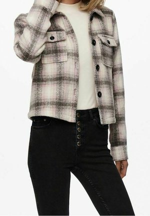 Kvinde iført en hvid top, ternet knapjakke i beige og brun samt højtsiddende sorte jeans med flere knapper foran.