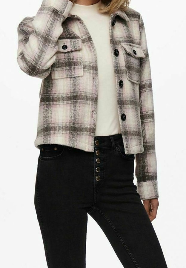 Femme portant un haut blanc, une veste à carreaux beige et marron boutonnée, et un jean noir taille haute avec plusieurs boutons à l'avant.