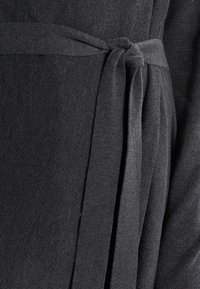 Prenda negra con una corbata de tela en la cintura, con una superficie suave y texturizada y elementos de diseño en capas. Construcción simple y elegante.