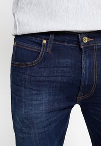Närbild av mörkblå denimbyxor med orange sömnad, kombinerat med en ljusgrå topp som är instoppad i midjan.