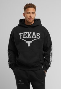 Crna dukserica s kapuljačom na kojoj je u velikim bijelim slovima ispisano "TEXAS", a ispod se nalazi grafika longhorna, s ispisom "LONGHORNS" u bijeloj boji na rukavu. Materijal je kombinacija pamuka.