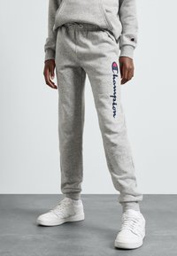 Pelēkas sporta bikses ar elastīgu jostasvīru, kurām sānos ir zilas krāsas "Champion" logo. Apvienotas ar baltām sneakerām. Strukturēta auduma.