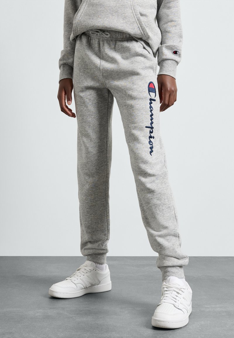 Pelēkas sporta bikses ar elastīgu jostasvīru, kurām sānos ir zilas krāsas "Champion" logo. Apvienotas ar baltām sneakerām. Strukturēta auduma.