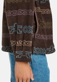 Manica di una giacca in pile marrone con motivi astratti colorati in blu, viola e beige, indossata sopra dei jeans in denim blu.