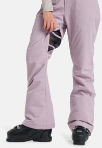 Licht paarse skibroek met een geventileerde ritszak, rechte pijpen, verstevigde boorden en gedragen met zwarte skischoenen met metalen hardware.