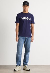 Jūras zila kokvilnas T-kreklā ar lielu balto "HUGO" logotipu, kombinācijā ar gaiši ziliem džinsiem un bēšiem sporta apaviem. Modelis arī nēsā melnu cepuri ar "HUGO".