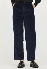 Pantalons larges en velours côtelé bleu marine avec une surface texturée en côtes, dotés d'une taille haute et de poches avant discrètes. Associés à des chaussures sombres.