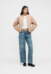 Ραφτό, ανοιχτό ροζ cropped μπουφάν με στρογγυλή λαιμόκοψη, συνδυασμένο με μπλε τζιν ψηλόμεσα παντελόνια με διακριτικό γραφικό μοτίβο. Μαύρα παπούτσια.