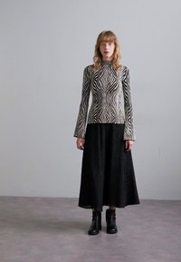 Top noir et crème à manches longues avec un motif en zigzag, associé à une jupe plissée noire et des bottines, se tenant sur un sol gris.
