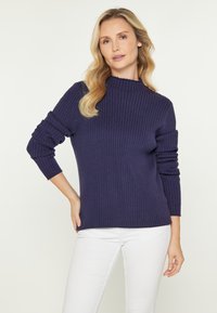 Marineblauer, gerippter Pullover mit hohem Kragen und langen Ärmeln, der einen lockeren Schnitt und strukturierte Maschen aufweist. Kombiniert mit weißen Hosen.