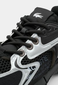 Zapatilla deportiva negra con un patrón de malla y acentos plateados. Los cordones son negros, con un logo en la lengüeta. Presenta ojetes de metal y una superficie texturizada.
