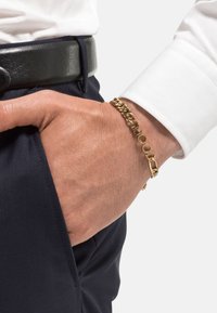 Pulsera de cadena de oro con eslabones circulares y un broche, llevada en una muñeca, combinada con una camisa blanca y pantalones oscuros.